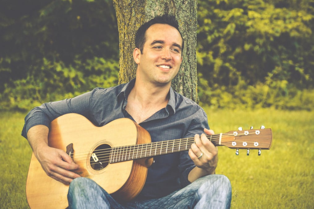 Austin MacRae sitting in front of a tree playing a guitar