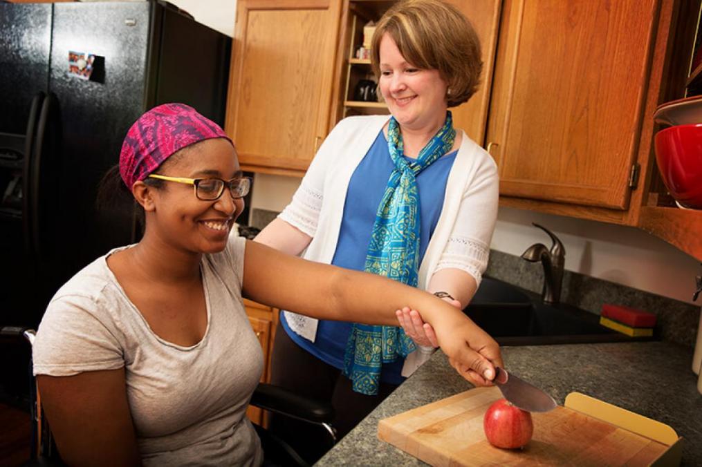 One woman sitting at a table and another woman next to her helping her cut an apple