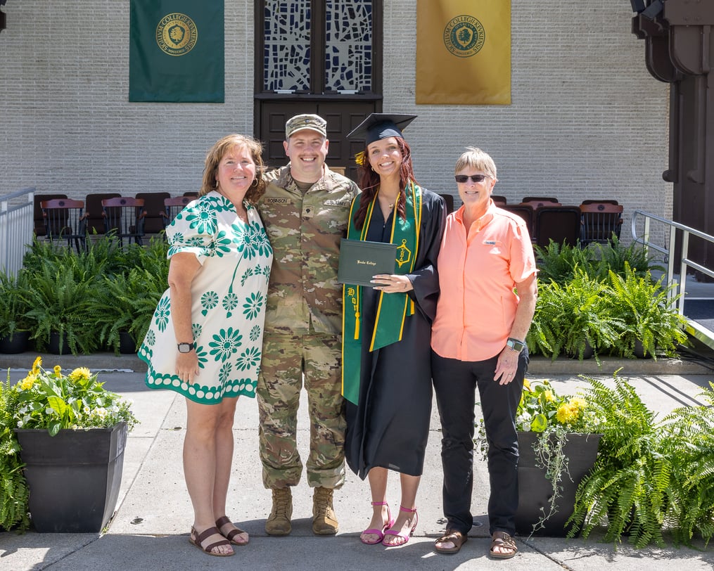 A graduate in cap and gown poses with family members and a person in military uniform while holding a diploma cover on the Keuka College Commencement stage.