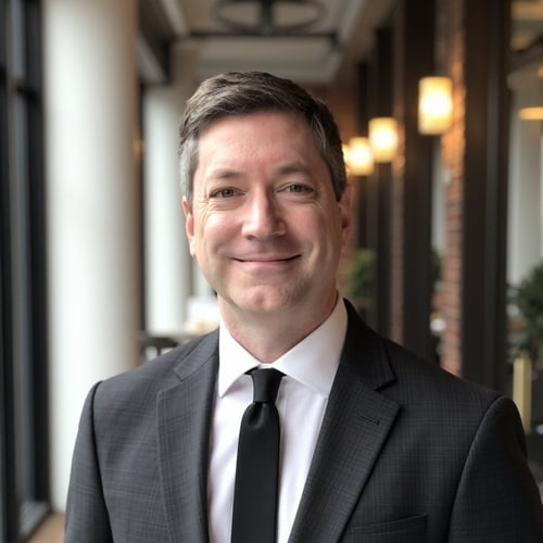 Middle-aged man in a dark suit and tie, smiling in a softly lit hallway with blurred lights in the background.