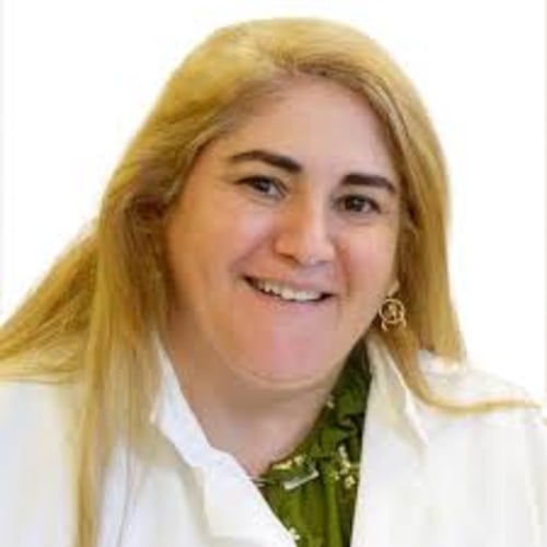 A close-up headshot of a smiling woman with long blonde hair, wearing a white top and a green patterned blouse underneath, against a plain light background.