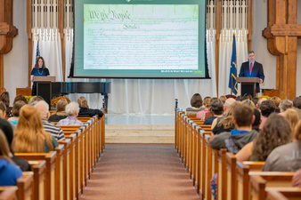 Two speakers address an audience from podiums in a chapel, with a projection screen displaying the U.S. Constitution behind them.
